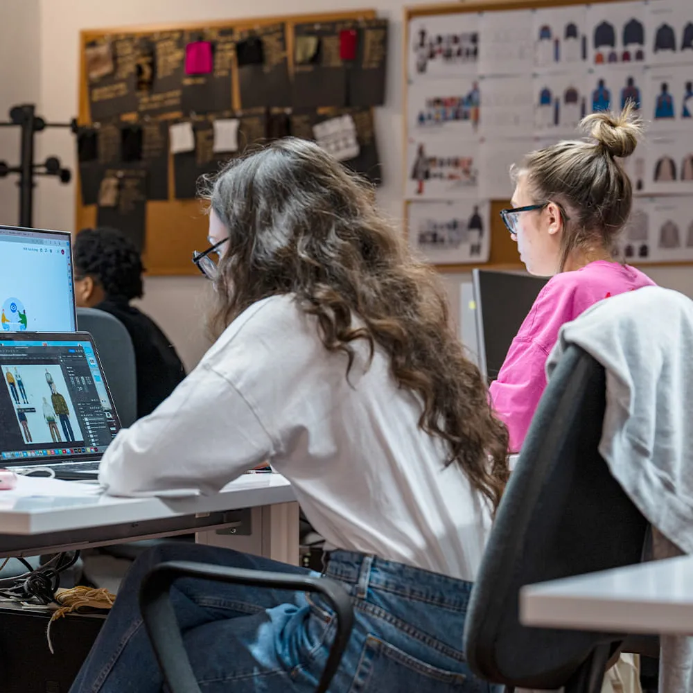 Due studentesse al computer in aula con in fondo una bacheca di figurini e schede tecniche